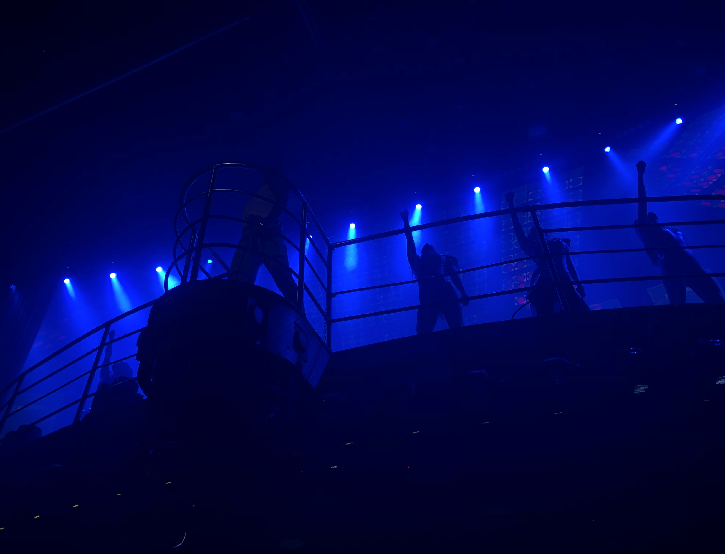From below shot of a stage with four performers, one featured on a curved, jutted out portion for the platform; the room is dark but the stage is illuminated in bright neon blue light and 3 of the background performers have their fists raised.